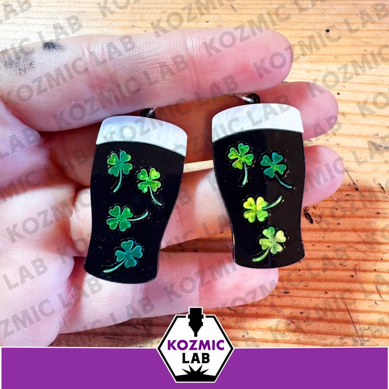 May include: A pair of black beer glass earrings with white foam tops and green shamrock designs. The earrings are held up against a wooden background. The earrings are a St. Patrick's Day theme.