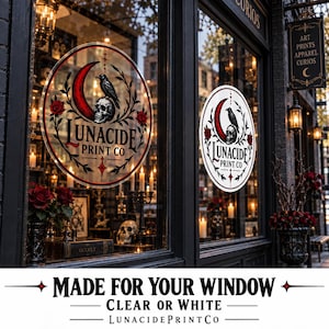 May include: A circular window decal featuring a red crescent moon, a black raven perched on a human skull, and red roses. The text reads LUNACIDE PRINT CO in a black serif font. The design is displayed on a glass storefront window.