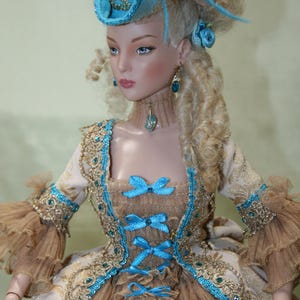 May include: A porcelain doll wearing a gold and blue gown with a blue feathered hat. The doll has blonde hair and blue eyes.