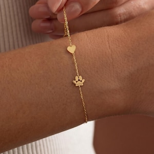 May include: A delicate gold bracelet featuring a heart charm and a paw print charm. The bracelet is worn on a wrist, showcasing the fine chain and the small, detailed charms. The paw print charm has the name "Aubrey" engraved on it.