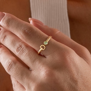 May include: A gold ring with the letter "D" and a small green gemstone. The ring is on a finger, and the design is delicate and minimalist.