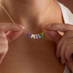 May include: A gold-colored necklace featuring the name "EMILY" spelled out in colorful, rounded letter beads. The letters are in purple, white, pink, blue, and green. The necklace is held up by a person.