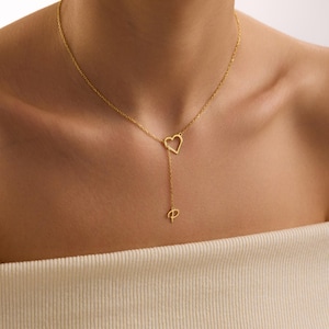 May include: A gold-coloured necklace featuring a heart-shaped pendant and a dangling charm. The necklace is worn around the neck, with the heart pendant positioned near the collarbone. The charm hangs below the heart. The necklace is made of a thin chain.