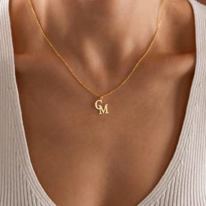 K&ouml;nnte beinhalten: Eine goldfarbene Halskette mit zarter Kette. Der Anh&auml;nger zeigt die verschlungenen Buchstaben "G" und "M". Die Halskette wird um den Hals getragen, der Anh&auml;nger liegt in der N&auml;he des Schl&uuml;sselbeins. Der Hintergrund ist eine neutrale Farbe.