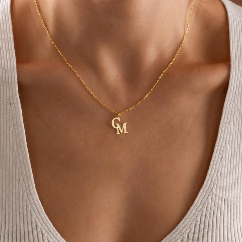 K&ouml;nnte beinhalten: Eine goldfarbene Halskette mit zarter Kette. Der Anh&auml;nger zeigt die verschlungenen Buchstaben "G" und "M". Die Halskette wird um den Hals getragen, der Anh&auml;nger liegt in der N&auml;he des Schl&uuml;sselbeins. Der Hintergrund ist eine neutrale Farbe.