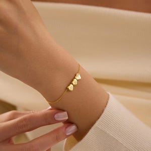 Puede incluir: Una delicada pulsera dorada con tres colgantes en forma de corazón. Cada colgante tiene una letra grabada. La pulsera se lleva en la muñeca, mostrando su fina cadena y los colgantes personalizados en forma de corazón.