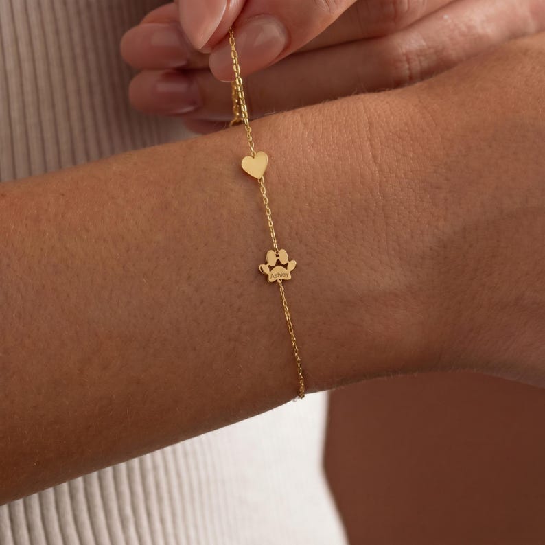 Puede incluir: Una delicada pulsera dorada con un dije de coraz&oacute;n y un dije de huella de pata. La pulsera se lleva en la mu&ntilde;eca, mostrando la fina cadena y los peque&ntilde;os y detallados dijes. El dije de huella de pata tiene grabado el nombre "Aubrey".
