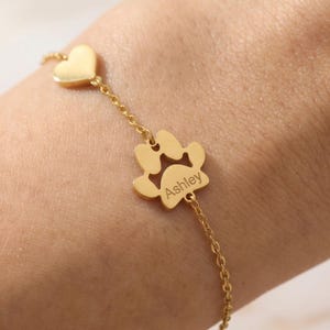 Puede incluir: Pulsera dorada con un dije de coraz&oacute;n y un dije de huella de pata con el nombre "Ashley" grabado. La pulsera de cadena fina es ideal para los amantes de las mascotas.