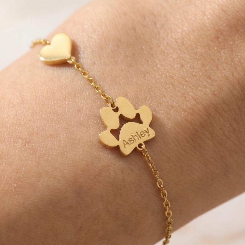Puede incluir: Pulsera dorada con un dije de coraz&oacute;n y un dije de huella de pata con el nombre "Ashley" grabado. La pulsera de cadena fina es ideal para los amantes de las mascotas.