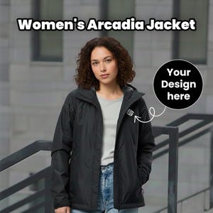 Pu&ograve; includere: Giacca Arcadia nera con cappuccio, indossata sopra una maglietta grigia e jeans blu. La giacca ha un piccolo logo sul petto e il testo "Women's Arcadia Jacket" &egrave; visualizzato in alto. &Egrave; presente anche un cerchio nero con "Your Design here".