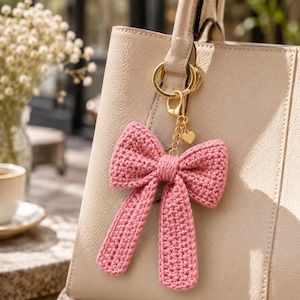 Puede incluir: Un llavero de lazo rosa tejido a ganchillo con un cierre dorado y un dije de corazón cuelga de un bolso beige. El lazo es de color rosa suave y tiene largas colas colgantes. El bolso es de color beige claro.