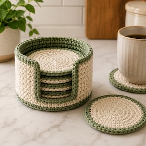 May include: A set of handmade crochet coasters in a holder. The coasters are off-white with a green border, and the holder is off-white with a green trim. A mug of coffee sits on a matching coaster. The coasters are approximately 4 inches in diameter.