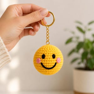 May include: A round, yellow crocheted keychain featuring a smiling face with black embroidered eyes and a curved mouth, along with two small pink stitched circles on the cheeks. The sphere is attached to a gold-toned metal chain and ring.