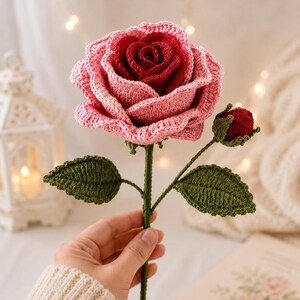 May include: A handmade crochet rose in shades of pink and red, with green leaves and stem. The flower is held in a hand, showcasing the intricate details of the yarn work. A bud is also present. The background is softly lit with a blurred effect.