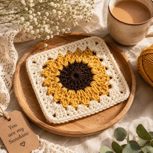 May include: A square, crocheted sunflower coaster with a brown center, yellow petals, and an off-white border, resting on a wooden plate. A tag with the text "You are my Sunshine" hangs nearby. A cup of coffee and yarn are also in the image.