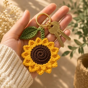 Pode incluir: Um porta-chaves de girassol em croché com um centro castanho escuro, pétalas amarelo-brilhante e uma única folha verde presa a um anel de metal dourado. Duas chaves de metal prateado estão presas ao anel, segurado numa mão.