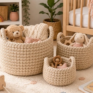 Puede incluir: Tres cestas de almacenamiento redondas, de color beige, tejidas a ganchillo a mano con asas integradas. La cesta más grande contiene un oso de peluche beige y un tejido color crema doblado. La mediana contiene un conejo de peluche color crema. La pequeña contiene juguetes de madera.
