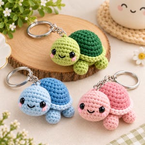 May include: Three crochet turtle keychains featuring small, round bodies with black bead eyes and pink stitched cheeks. One turtle has a light green head and dark green shell, one is light blue, and one is light pink, each with a metal ring.