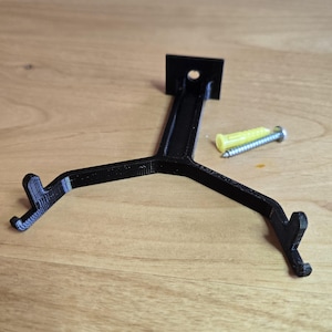 May include: A black Y-shaped wall mount bracket with two upward-facing hooks at the ends of the arms and a single screw hole at the top base. The bracket is accompanied by a yellow plastic wall anchor and a silver metal screw for installation.