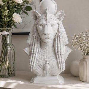 May include: A white matte bust sculpture of a lioness deity featuring a sun disk headdress, braided hair, ankh earrings, and a central scarab beetle pendant on the chest. The figure is depicted with detailed facial features and ornate collar.