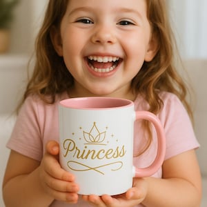 Op de afbeelding: Een witte keramische mok met een roze handvat en rand, met het woord "Princess" in gouden letters, vastgehouden door een lachend kind. De mok heeft een kroonontwerp boven het woord. Het kind draagt een roze shirt.