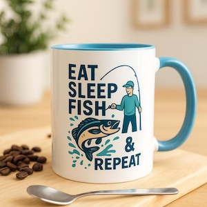 Könnte beinhalten: Weißer Keramikbecher mit blauem Innenraum und Henkel. Der Becher zeigt den Text "EAT SLEEP FISH & REPEAT" mit einer Grafik eines Anglers und eines Fisches. Ein Löffel und Kaffeebohnen liegen auf einer Holzoberfläche.