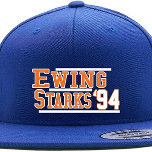 May include: Royal blue snapback hat with the text "EWING STARKS '94" in orange and white. The hat has a flat brim and a green underside.