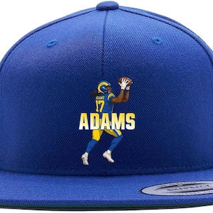 May include: A royal blue snapback hat featuring a graphic of a football player catching a ball. The player is wearing a yellow and blue uniform with the name "ADAMS" in white block letters. The hat has a flat brim.