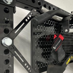 Pode incluir: Equipamento de halterofilismo preto montado em um rack de metal preto. Inclui uma barra de pull-up, vários acessórios de aderência e cintas de halterofilismo. O rack tem marcações numeradas para ajustes. O equipamento é projetado para exercícios de treinamento de força.