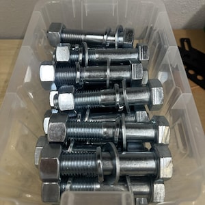 May include: A collection of silver-colored metal bolts, each featuring a hexagonal head, a threaded shaft, a flat washer, and a hexagonal nut, all contained within a clear, rectangular plastic storage bin with a visible grid-like structure.