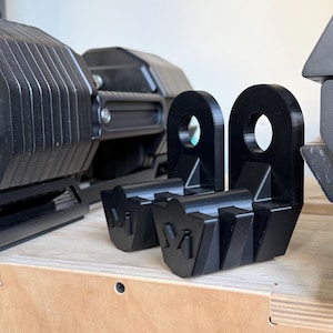 Może przedstawiać: Czarne metalowe wyposażenie do podnoszenia ciężarów, z dwoma wspornikami z pętlą i rowkową podstawą. Na podstawie wytłoczona jest litera "M". Inny sprzęt do podnoszenia ciężarów jest widoczny w tle.