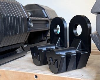 May include: Two black 3D-printed plastic attachments with a circular hole at the top and a shield-shaped base featuring a raised letter M. These components are positioned in front of a larger black dumbbell-style weight with a textured surface.