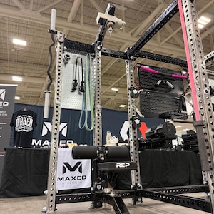 May include: A black and silver steel power rack with a REP branded adjustable bench, featuring a vertical pegboard attachment, a barbell holder, and a pulley system. The structure includes numbered uprights and various gym accessories.