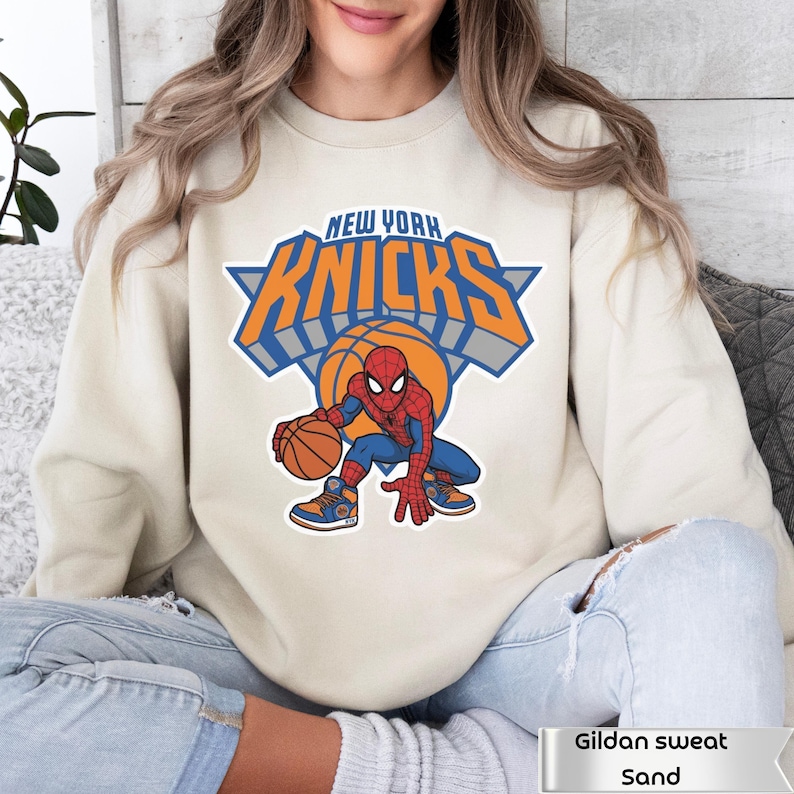 May include: A sand-colored crewneck sweatshirt featuring a graphic print of the New York Knicks logo with the text "NEW YORK KNICKS" in orange and blue. Below the logo, a cartoon Spider-Man character is shown dribbling a basketball while wearing sneakers.