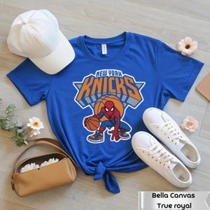 May include: A royal blue short-sleeve t-shirt featuring a graphic of Spider-Man holding a basketball with the text New York Knicks above it. The shirt is displayed with a white baseball cap, white sneakers, and a tan handbag.