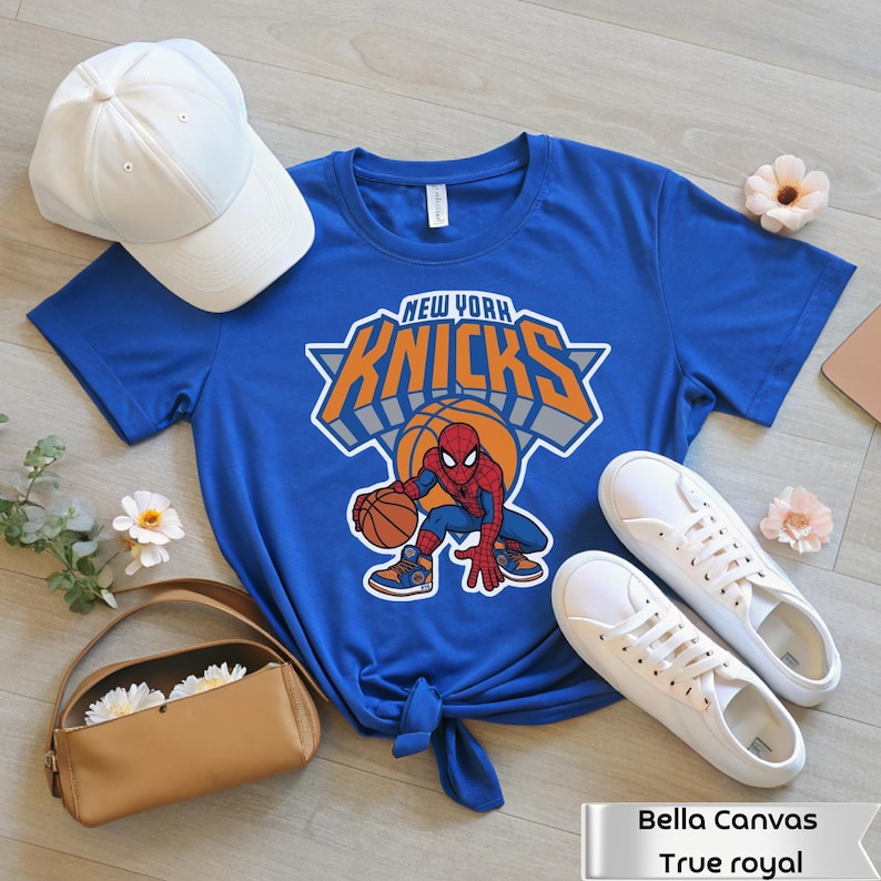 May include: A royal blue short-sleeve t-shirt featuring a graphic of Spider-Man holding a basketball with the text New York Knicks above it. The shirt is displayed with a white baseball cap, white sneakers, and a tan handbag.