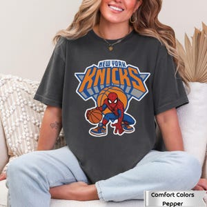 May include: A charcoal gray short-sleeve t-shirt featuring a graphic of the Marvel character Spider-Man dribbling a basketball. Above the character, the text reads NEW YORK KNICKS in orange and blue block letters with a white outline.