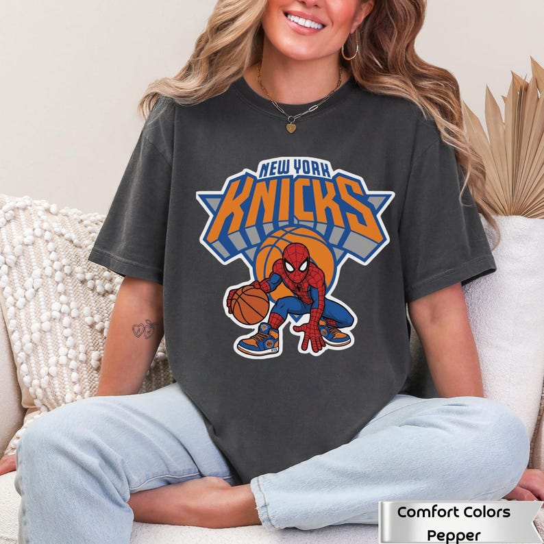 May include: A charcoal gray short-sleeve t-shirt featuring a graphic of the Marvel character Spider-Man dribbling a basketball. Above the character, the text reads NEW YORK KNICKS in orange and blue block letters with a white outline.