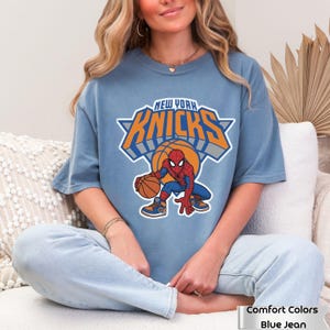 May include: A dusty blue short-sleeve t-shirt featuring a graphic of the New York Knicks logo with the text "NEW YORK KNICKS" in orange and blue, positioned above a cartoon illustration of Spider-Man dribbling a basketball while wearing sneakers.