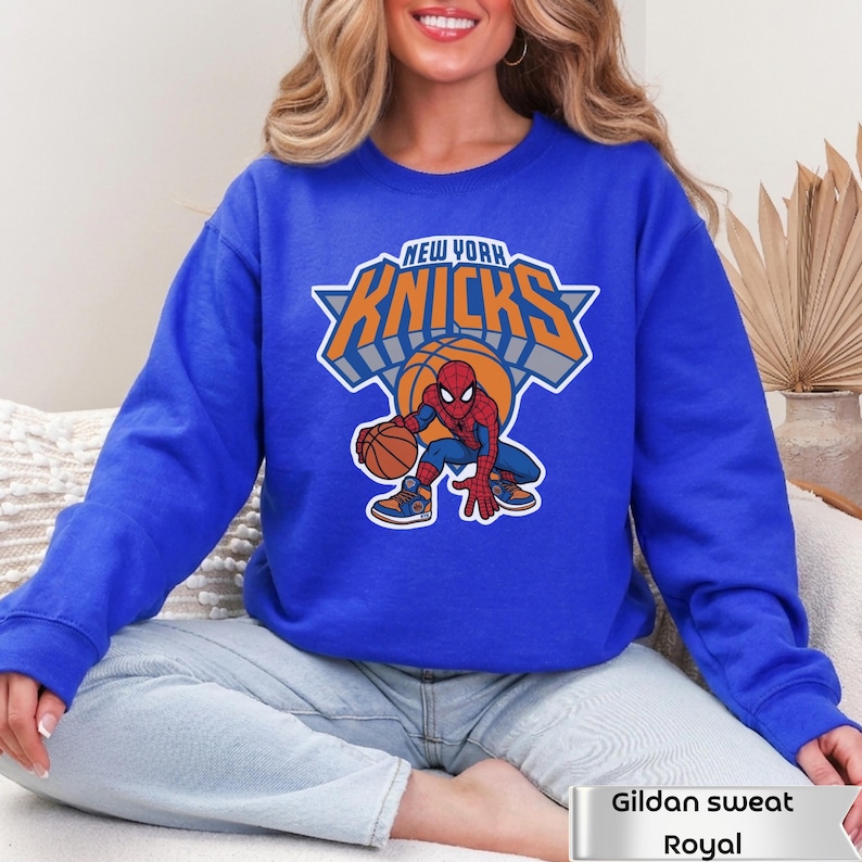 May include: A royal blue crewneck sweatshirt featuring a graphic of the New York Knicks logo with the text NEW YORK KNICKS in orange and white, and a cartoon illustration of Spider-Man dribbling a basketball in the center of the design.