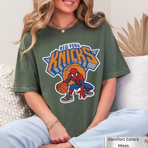 May include: A moss green short-sleeve t-shirt featuring a graphic of the New York Knicks logo with a basketball and Spider-Man character. The text reads New York Knicks. The shirt is labeled as Comfort Colors Moss.