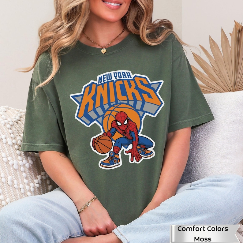 May include: A moss green short-sleeve t-shirt featuring a graphic of the New York Knicks logo with a basketball and Spider-Man character. The text reads New York Knicks. The shirt is labeled as Comfort Colors Moss.