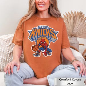 May include: A burnt orange t-shirt featuring a graphic of the New York Knicks logo with the text "NEW YORK KNICKS" above a basketball and a cartoon Spider-Man character dribbling a basketball. The shirt includes a label reading "Comfort Colors Yam".