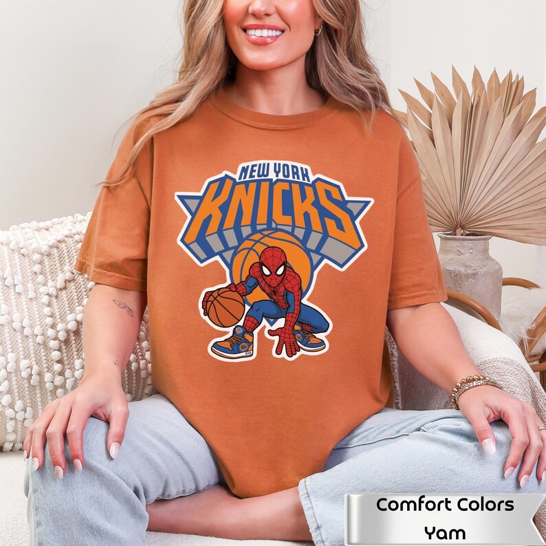 May include: A burnt orange t-shirt featuring a graphic of the New York Knicks logo with the text "NEW YORK KNICKS" above a basketball and a cartoon Spider-Man character dribbling a basketball. The shirt includes a label reading "Comfort Colors Yam".