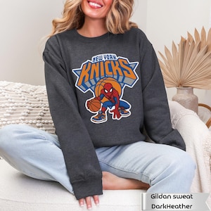 May include: A dark heather gray crewneck sweatshirt featuring a graphic of the Marvel character Spider-Man dribbling a basketball. Above the character, the text reads NEW YORK KNICKS in bold orange and blue lettering with a white outline.