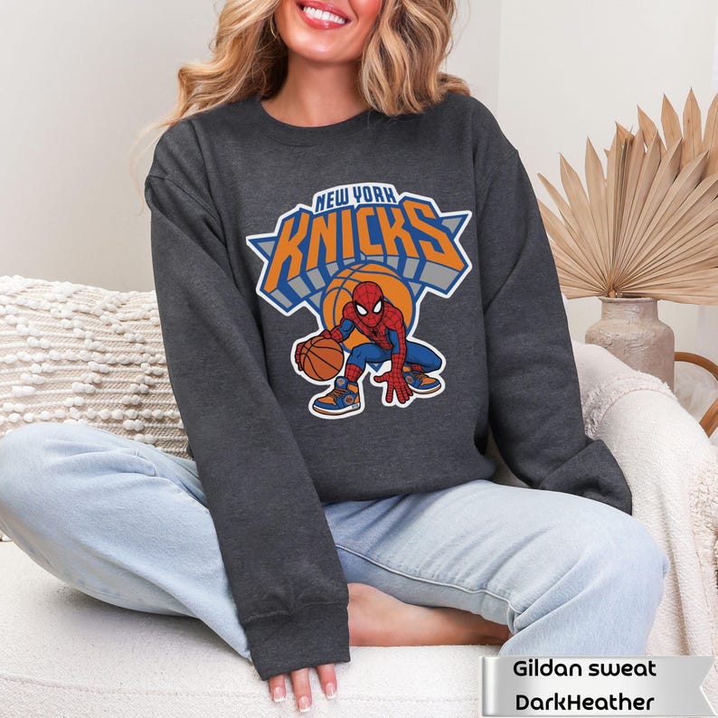 May include: A dark heather gray crewneck sweatshirt featuring a graphic of the Marvel character Spider-Man dribbling a basketball. Above the character, the text reads NEW YORK KNICKS in bold orange and blue lettering with a white outline.