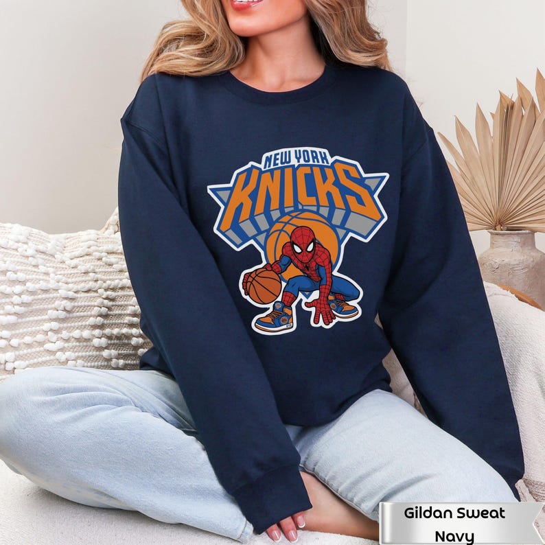 May include: A navy blue crewneck sweatshirt featuring a graphic of the Marvel character Spider-Man dribbling a basketball. Above the character, the text reads NEW YORK KNICKS in orange and white lettering with a grey shadow effect.