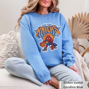 May include: A light blue crewneck sweatshirt featuring a graphic of the New York Knicks logo with the text "NEW YORK KNICKS" in orange and grey, and a cartoon illustration of Spider-Man dribbling a basketball in the center of the design.