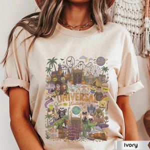 May include: An ivory t-shirt featuring a colorful graphic print of Universal Studios characters and attractions, including a roller coaster, palm trees, and various animated figures. The text Universal Studios is printed in the center of the design.