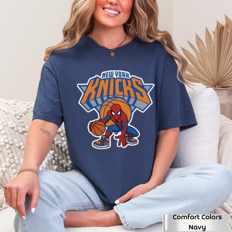 May include: A navy blue short-sleeve t-shirt featuring a graphic of the Marvel character Spider-Man dribbling a basketball. Above the character, the text reads NEW YORK KNICKS in orange and white lettering with a grey drop shadow effect.